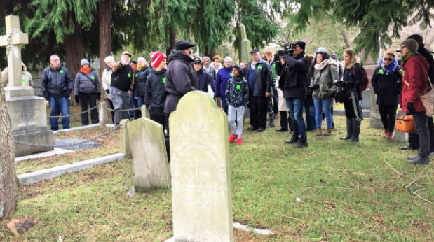 Group of about 30 people, women, men and youth, wearing coats/hats gathered around gravesites at Ross Bay Cemetery, speaker stands in front of one of the grave markers, grass underfoot, trees in background