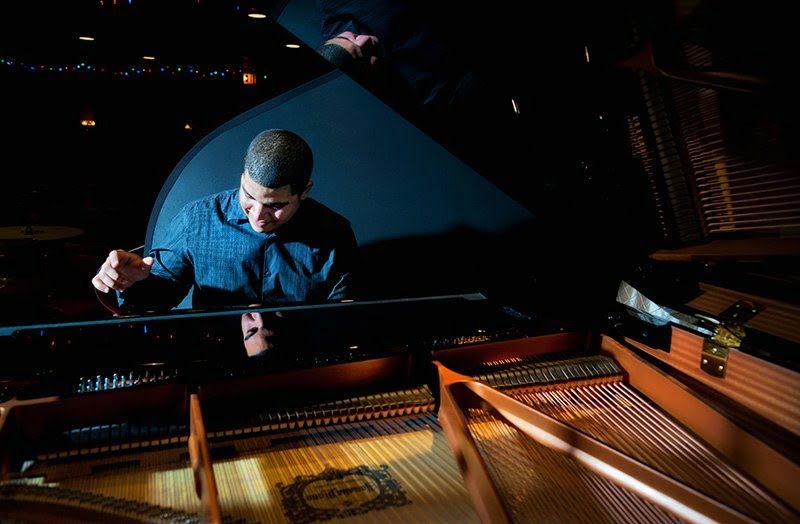 black man in blue checked shirt playing piano, spotlights in background, piano with open lid in foreground